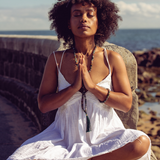 Woman sitting with beach in the background. Her eyes are closed and her hands in prayer position and she is wearing Ambarya Good health - Bloodstone Mala Bead Set